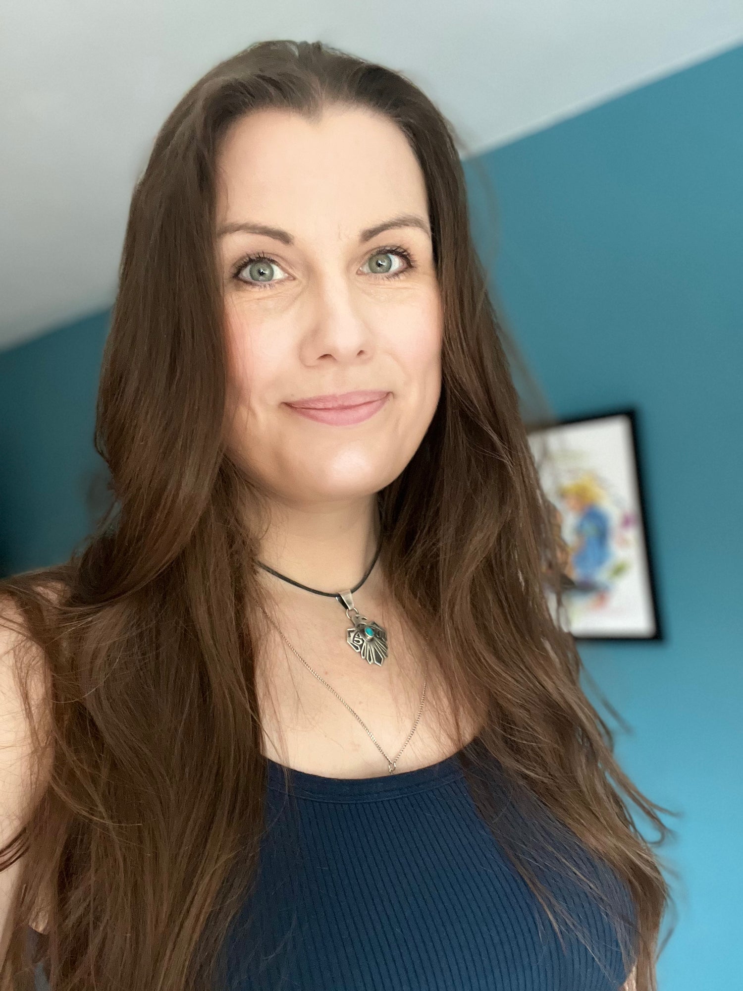 Woman with long brown hair wearing a blue top and necklace in a room with a blue wall.